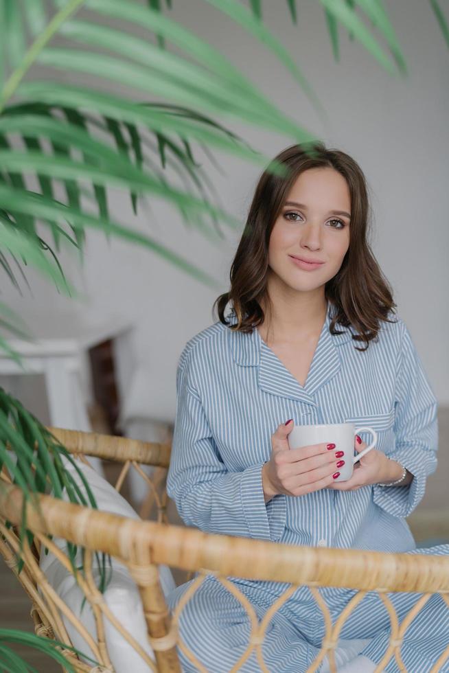 Adorable young female has charming look, dressed in striped domestic costume, drinks hot beverage, sits in wicker chair, enjoys rest and day off. People, lifestyle and recreation time concept Stock Free