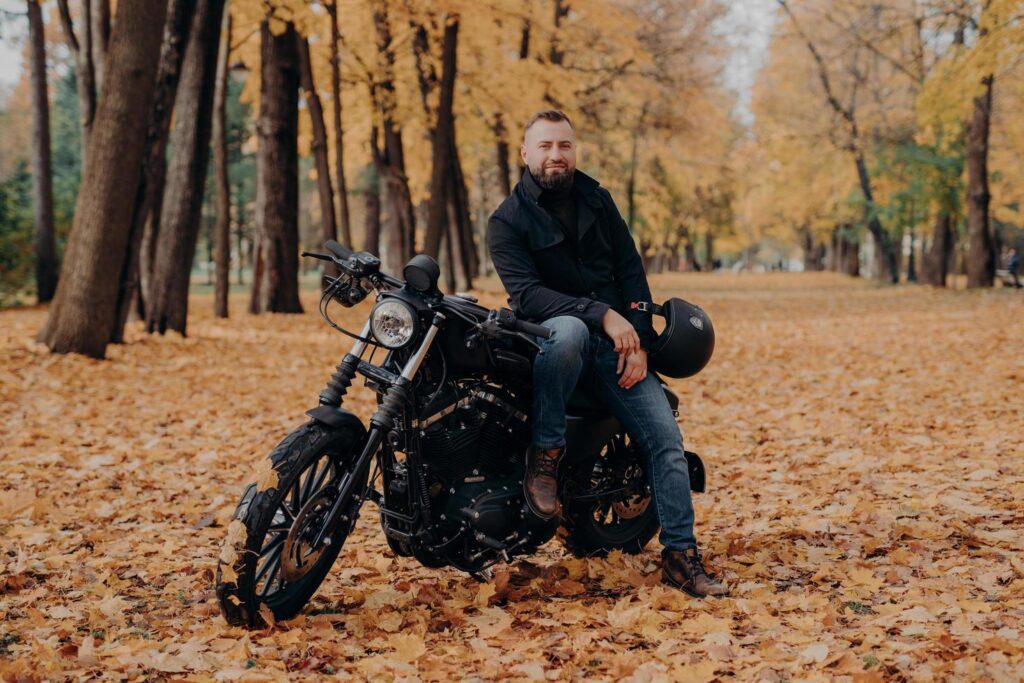 Bearded biker poses on own motorcycle, holds helmet, rides motorbike, poses outdoor in park, ground covered with fallen leaves. Biker lifestyle. Adventure on own transport. Male rider outdoor Stock Free