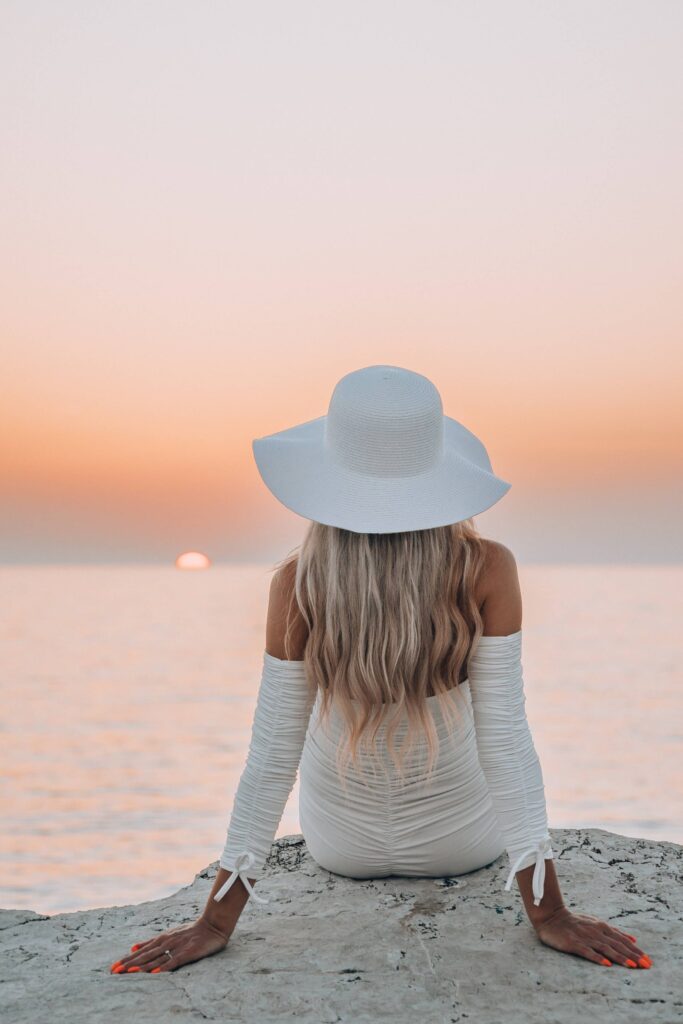 Beautiful Woman in White Hat Sitting on a Rock and Enjoying Sea Sunset Free Photo