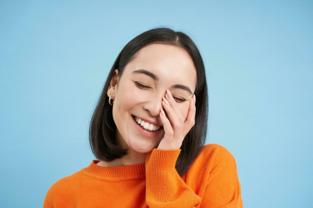 Close up of beatiful brunette woman, laughing and smiling, touches her face, expresses pure joy, stands over blue background Stock Free