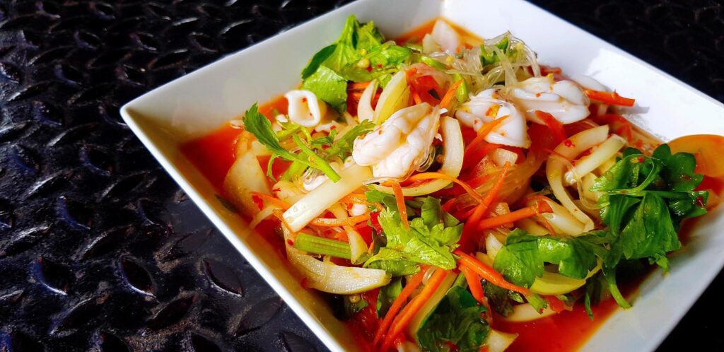 Close up Thai spicy seafood salad with glass noodle, squid, shrimp, sliced tomato, onion, carrot and celery on white dish or plate. Asian and famous food on black stainless steel background. Stock Free