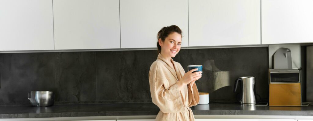 Daily routine and lifestyle. Young beautiful woman in bathrobe, standing in kitchen with cup of coffee, drinking tea, smiling and looking happy Stock Free