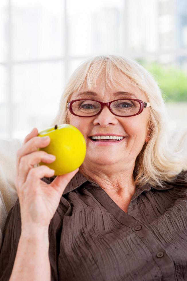 Enjoying healthy lifestyle. Cheerful senior woman holding apple and looking at camera while sitting at the chair Stock Free