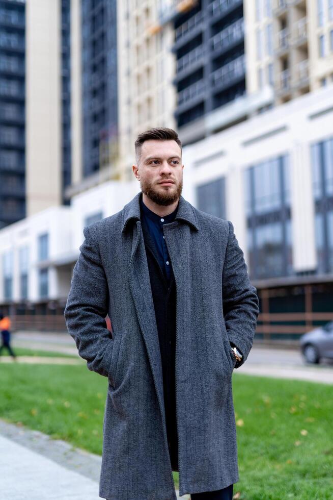 Fashionable man in coat stands near office centre and watching forward. Business person keeps calm and thinking. Successful concept. Thoughtful and resolute look of handsome man in big town. Stock Free