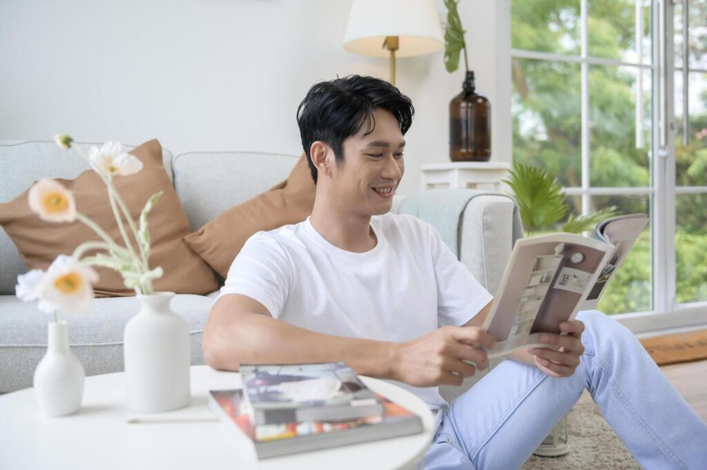 Happy asian man sitting on couch reading a book in living room at home, relax time and lifestyle concept Stock Free