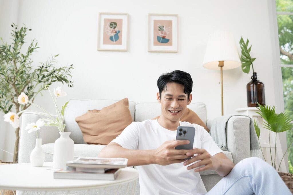 Happy asian man sitting on couch using smartphone in living room at home, relax time and lifestyle concept Stock Free