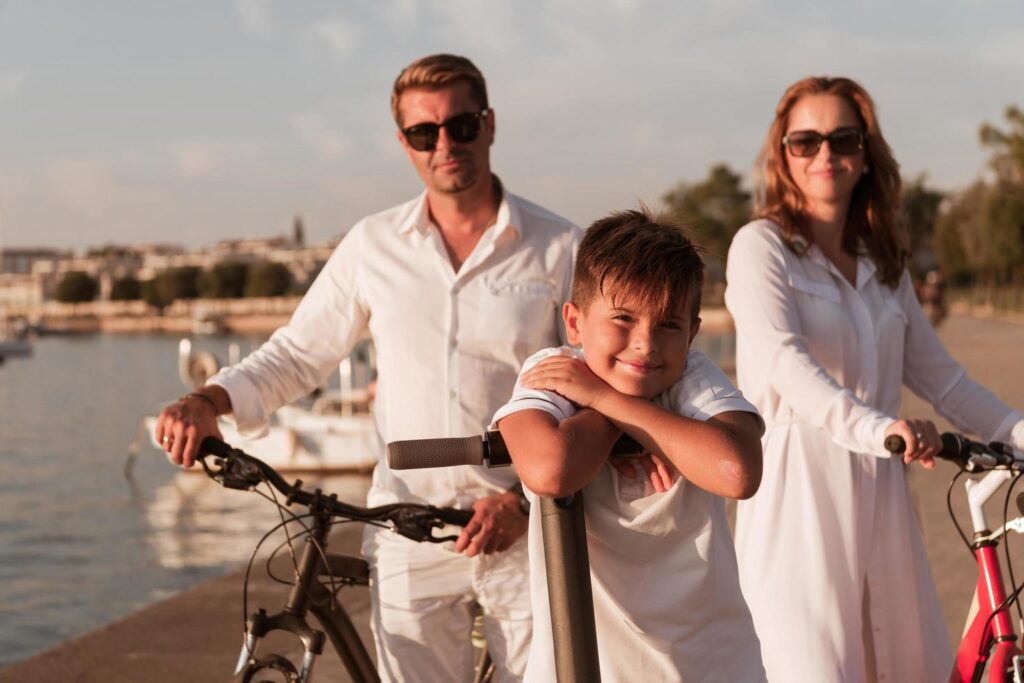 Happy family enjoying a beautiful morning by the sea together, parents riding a bike and their son riding an electric scooter. Selective focus Stock Free