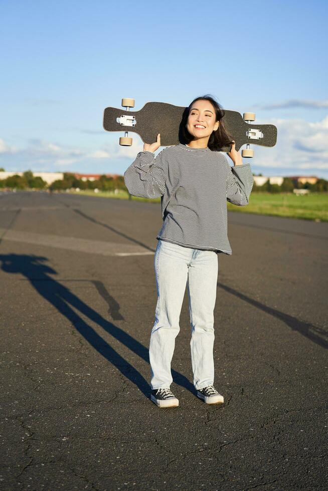 Lifestyle and people. Young asian girl posing with longboard, skating on her cruiser. Smiling woman holding skateboard on shoulders Stock Free