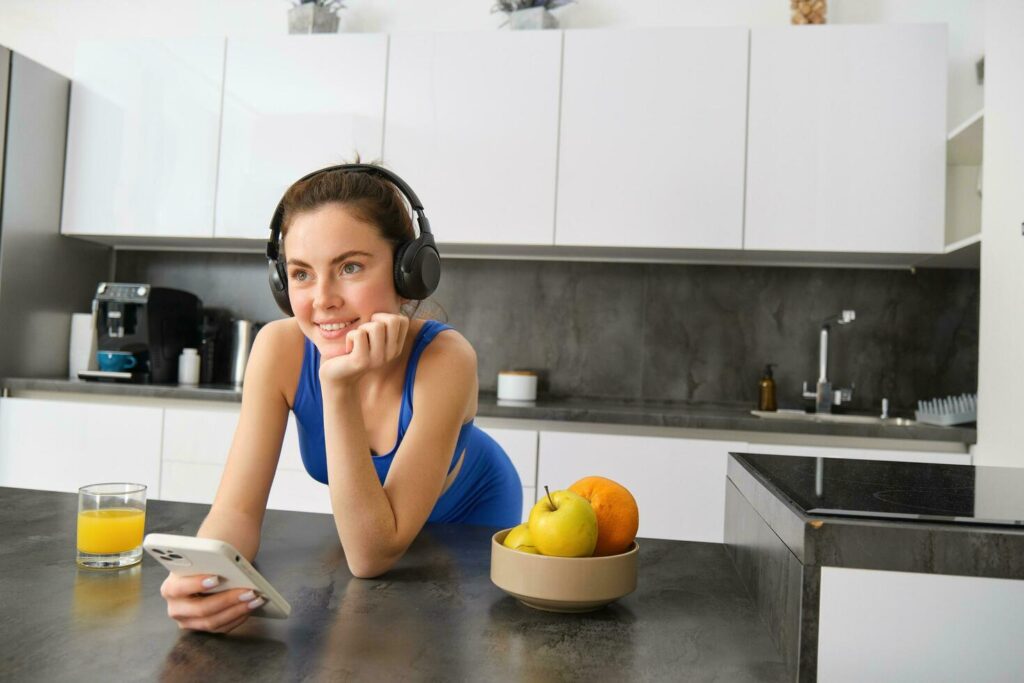 Lifestyle and workout. Young smiling woman in headphones, standing in kitchen with smartphone, drinking orange juice and listening music, heading over to gym, going jogging Stock Free