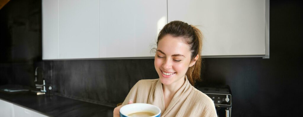 Lifestyle concept. Portrait of happy brunette woman in bathrobe, drinking coffee in the kitchen, having morning cuppa and smiling Stock Free