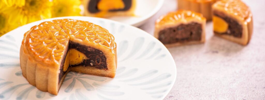 Mid-Autumn Festival traditional food concept – Beautiful cut moon cake on blue pattern plate on white background with flower, close up, copy space Stock Free