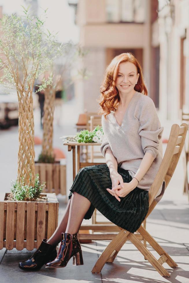 Photo of beautiful red haired woman dressed in stylish clothes, sits in open air, poses at cafeteria, has pleasant appearance, enjoys leisure time, smiles positively. People and lifestyle concept Stock Free