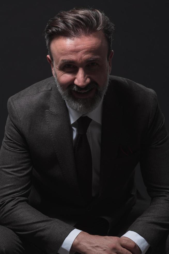 Portrait of adult businessman wearing trendy suit and sitting in modern studio on stylish chair against the black background Stock Free