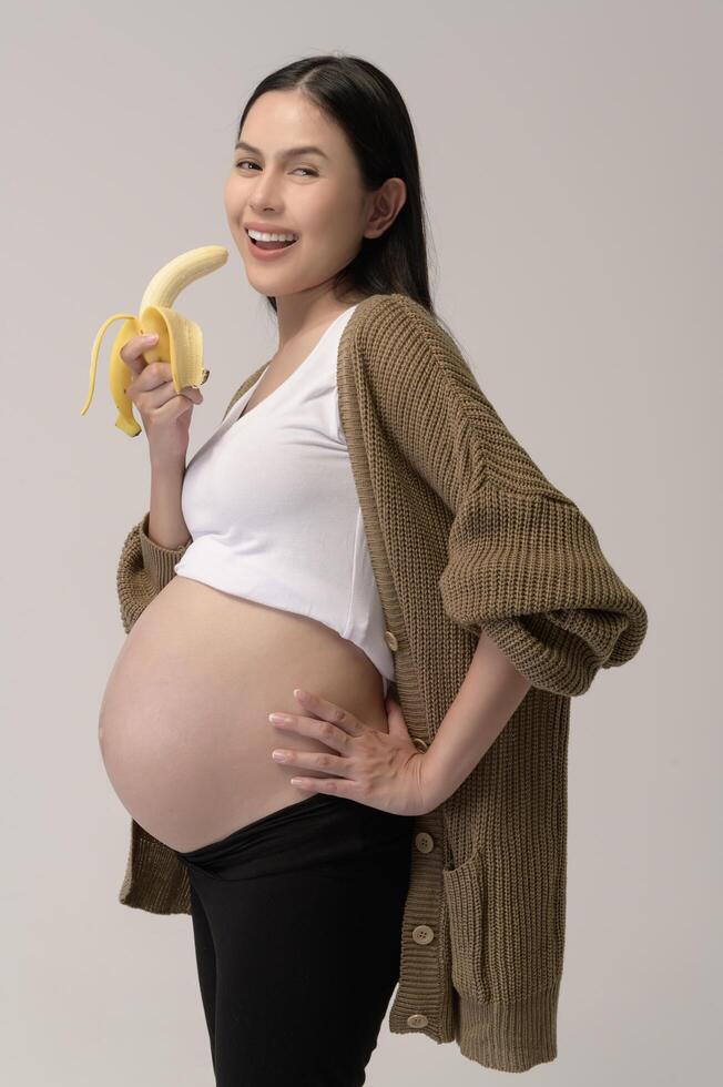 Portrait of Beautiful pregnant woman holding banana over white background studio, health and maternity concept Stock Free