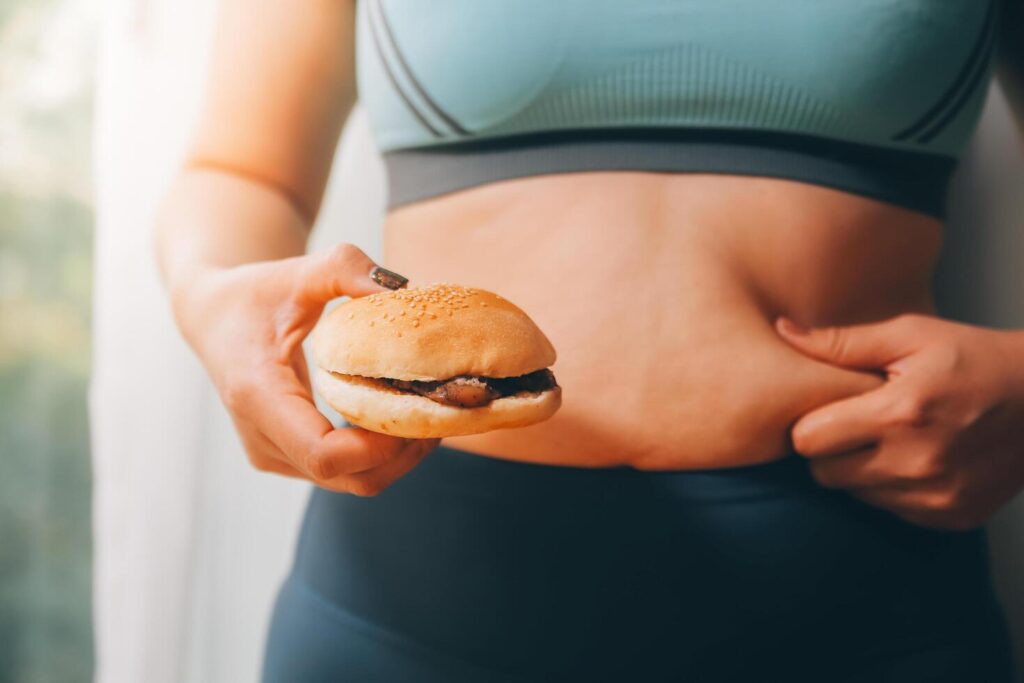 Portrait of obese woman looks sad while pinching her belly fat surrounded by junk foods. Diet fail concept Stock Free