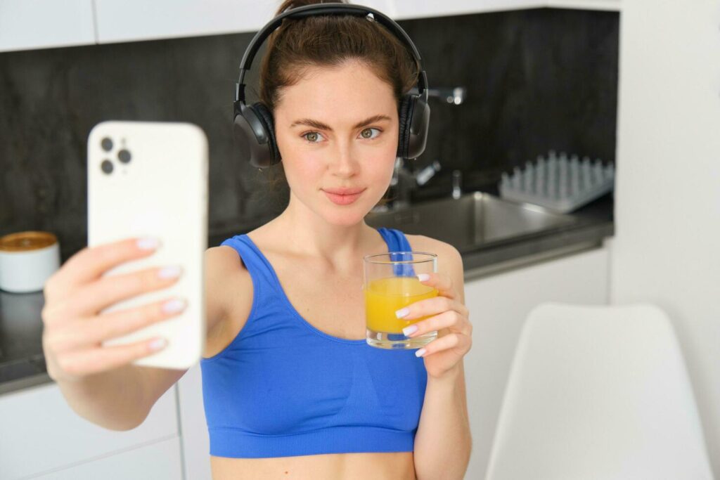 Portrait of young lifestyle, fitness blogger, woman with orange juice, takes selfie on smartphone camera, posing in activewear and wireless headphones Stock Free