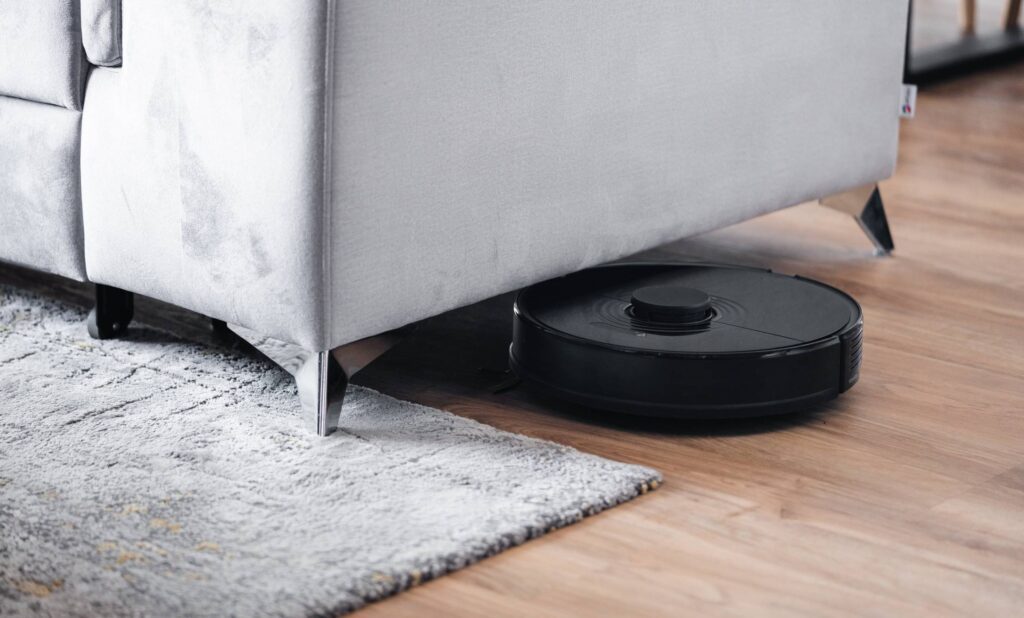 Robotic Vacuum Cleaner Cleaning Floor Around Gray Sofa Free Photo