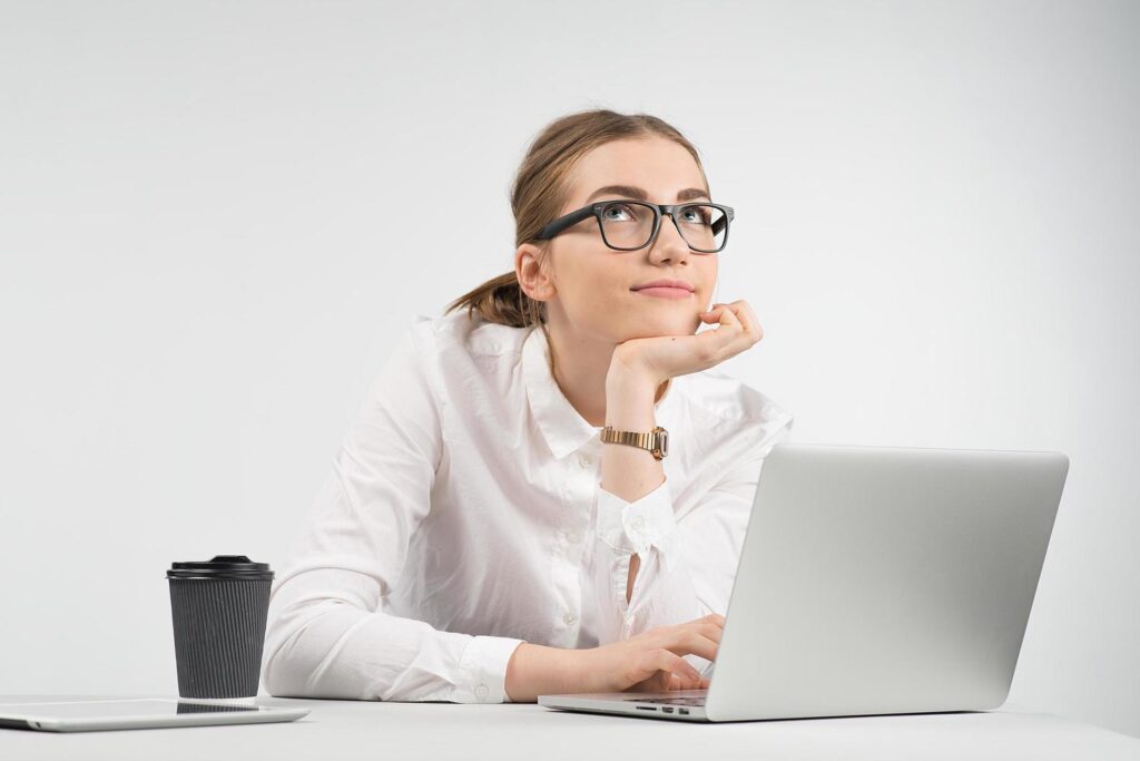 Smiling business woman sitting behind a laptop with a cup of coffee and ipad on the table and looking up dreamy Stock Free