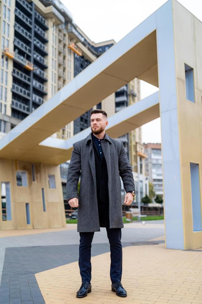 Street business style. Man in an elegant dark suit and gray coat. Handsome young caucasian man is standing on a central street with up to date architecture. Stock Free