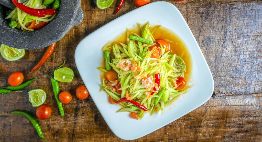 Thai food papaya salad on a plate with lemon and tomato on a wooden background.Top view Stock Free