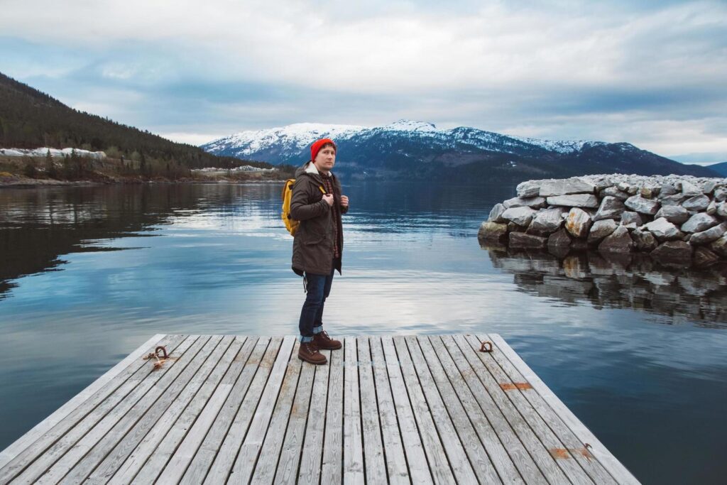 Traveler man with a yellow backpack wearing a red hat standing on the background of mountain and lake wooden pier. Travel lifestyle concept Stock Free