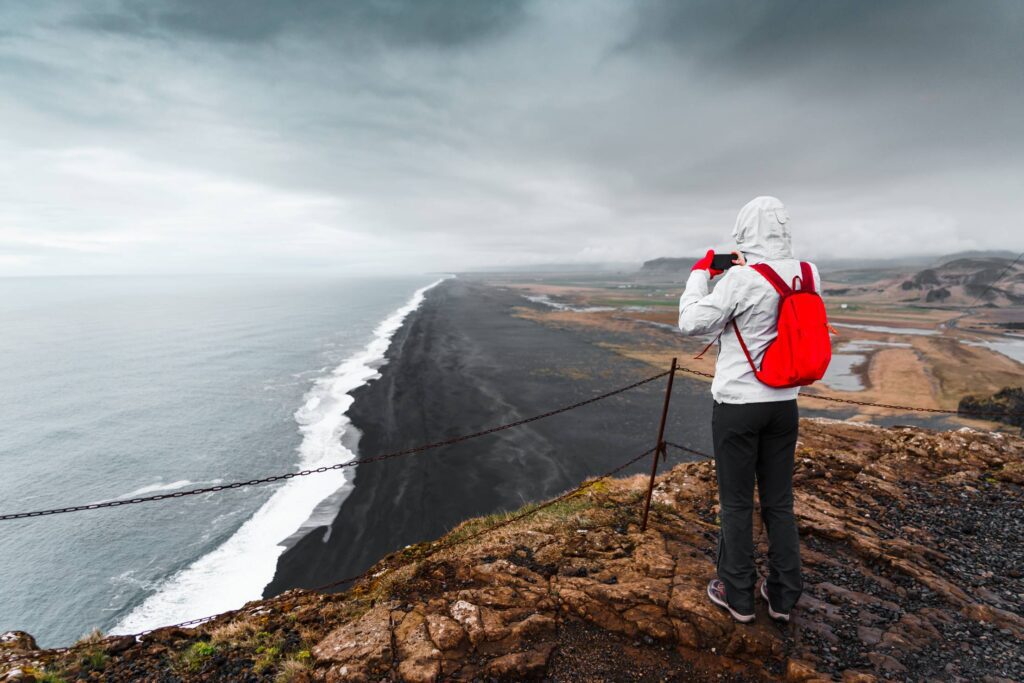 Woman Traveler Standing on Top of a Rock and Taking Pictures of a Black Beach in Iceland Free Photo