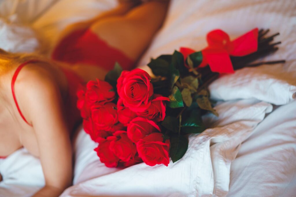 Woman Wearing Red Lingerie Lying in Bed with a Bouquet of Red Roses Free Photo