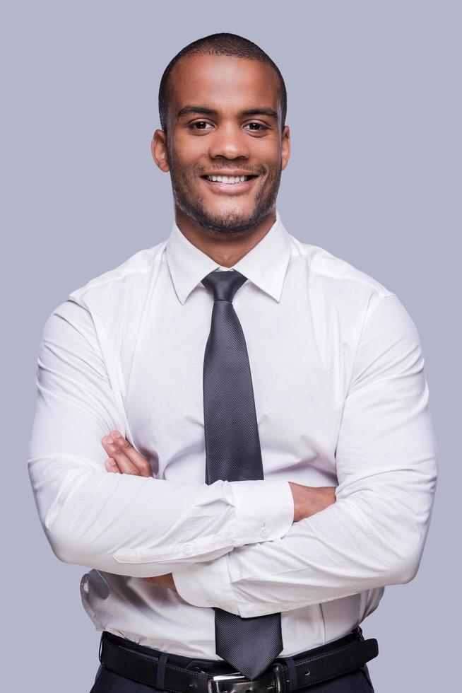 Young and successful. Confident young African man in shirt and tie keeping arms crossed and smiling while standing against grey background Stock Free