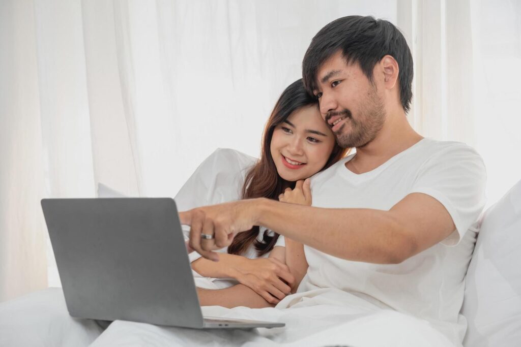 Young happy asian couple using laptop in bed watching movie, romantic time to enhance family relationship, family concept. Stock Free