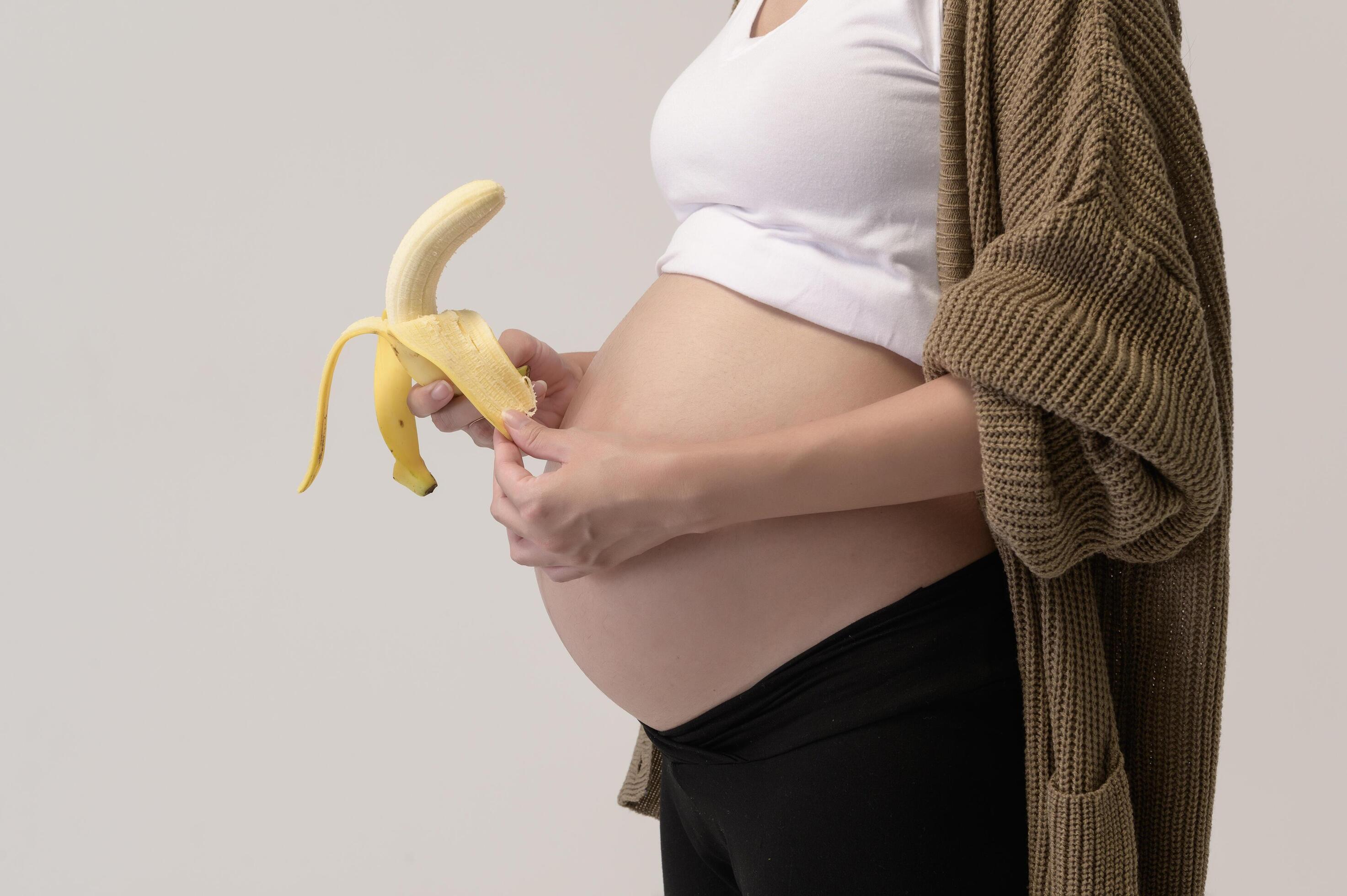 Portrait of Beautiful pregnant woman holding banana over white background studio, health and maternity concept Stock Free