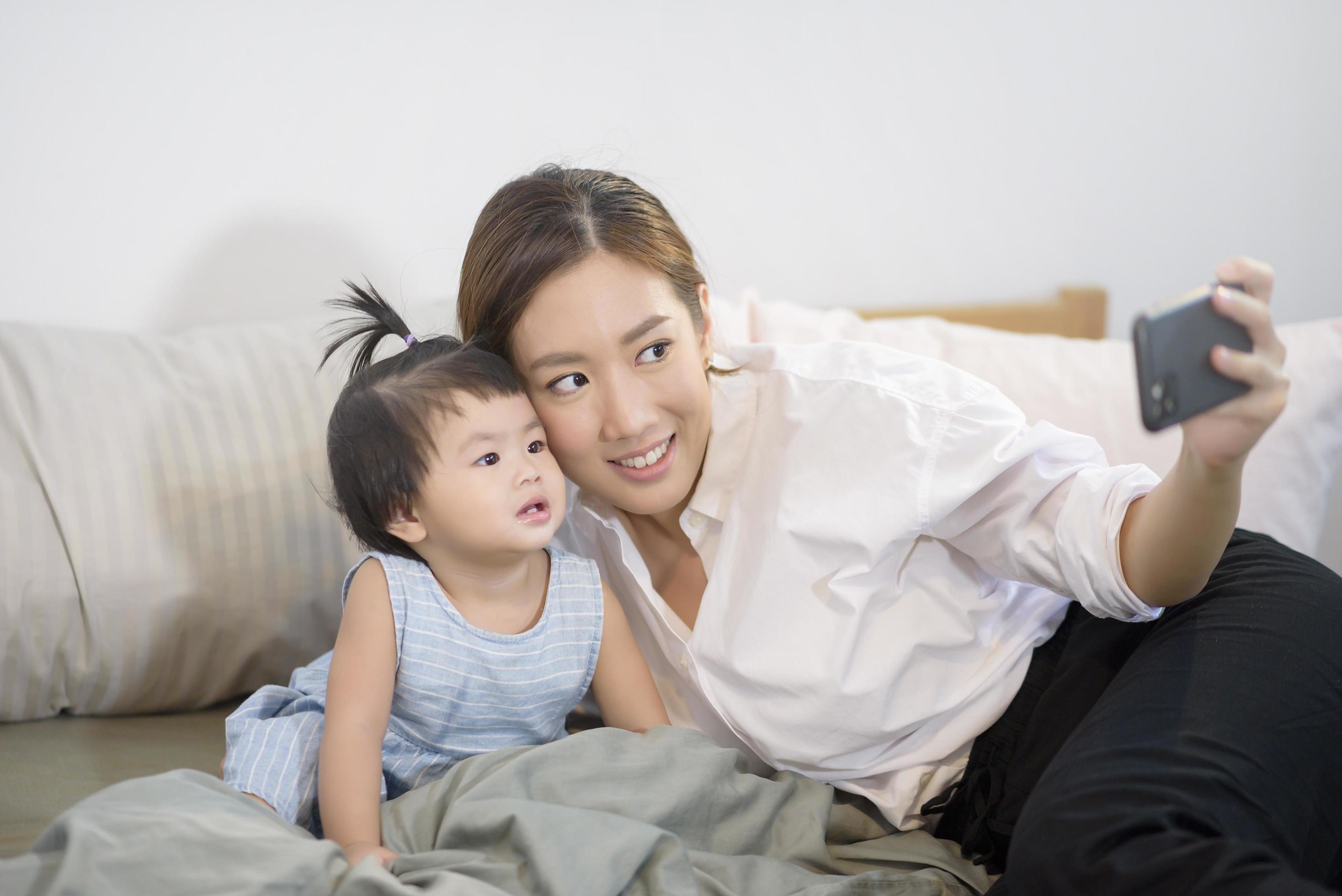 Asian Mother and her baby daughter are making selfie or video call to father in bed , Family , Home safety , parenthood , technology concept Stock Free