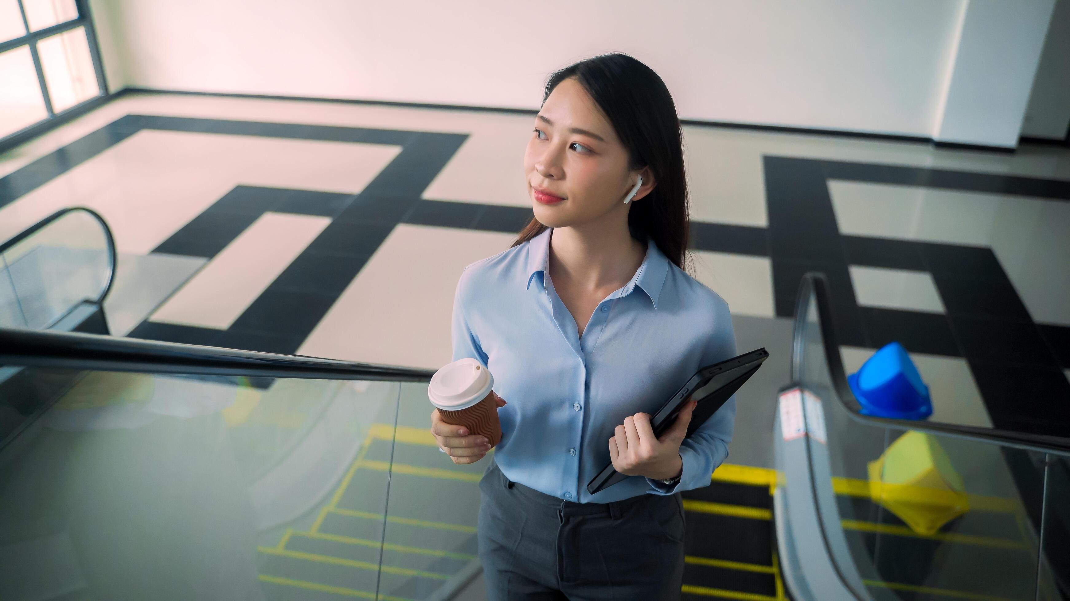 A business woman is walking up to escalator with a coffee cup in her hand Stock Free