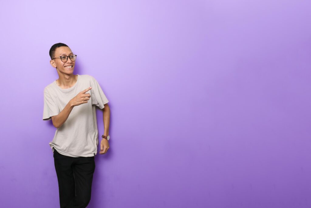 an Asian man in a plain white t-shirt with glasses gesturing to something to the side isolated on purple background Stock Free