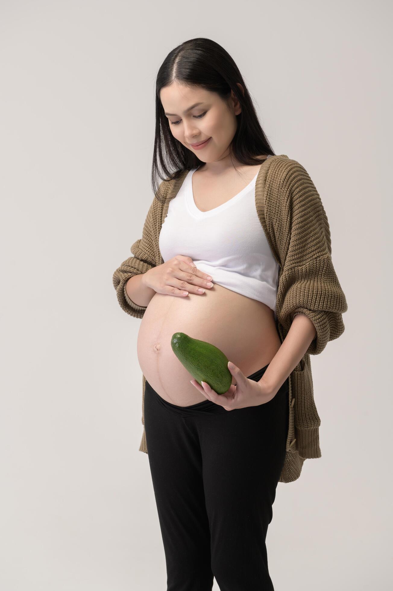 Portrait of Beautiful pregnant woman holding avocado over white background studio, health and maternity concept Stock Free