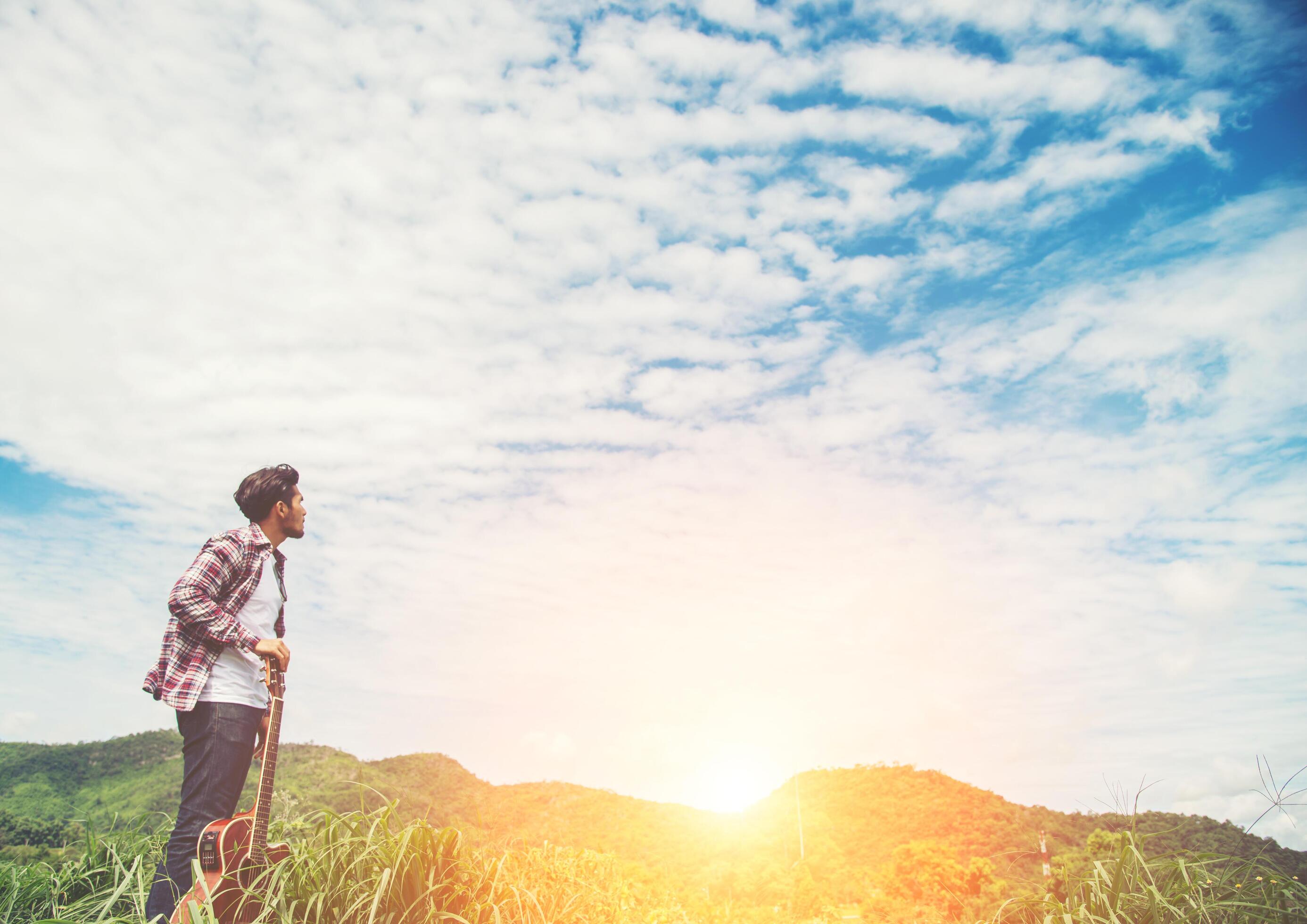 Young hipster man holding a guitar with a walking in nature, Relaxing in the field in a sunny blue sky day. Stock Free