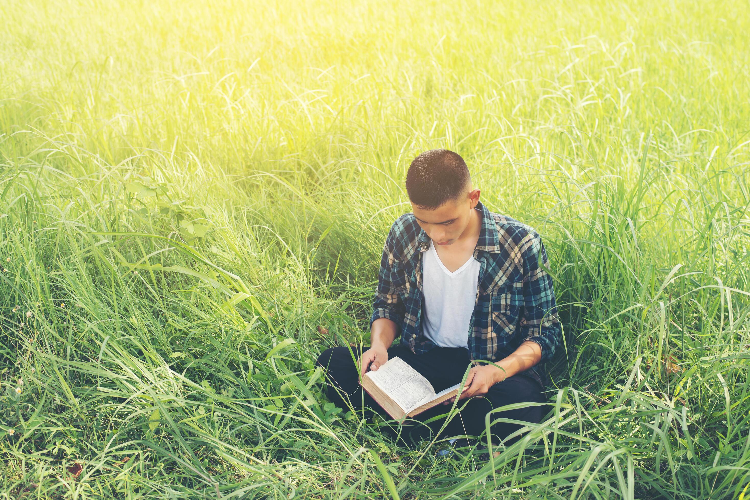 Young hipster man sitting on grassland reading book with nature around so happy. Stock Free