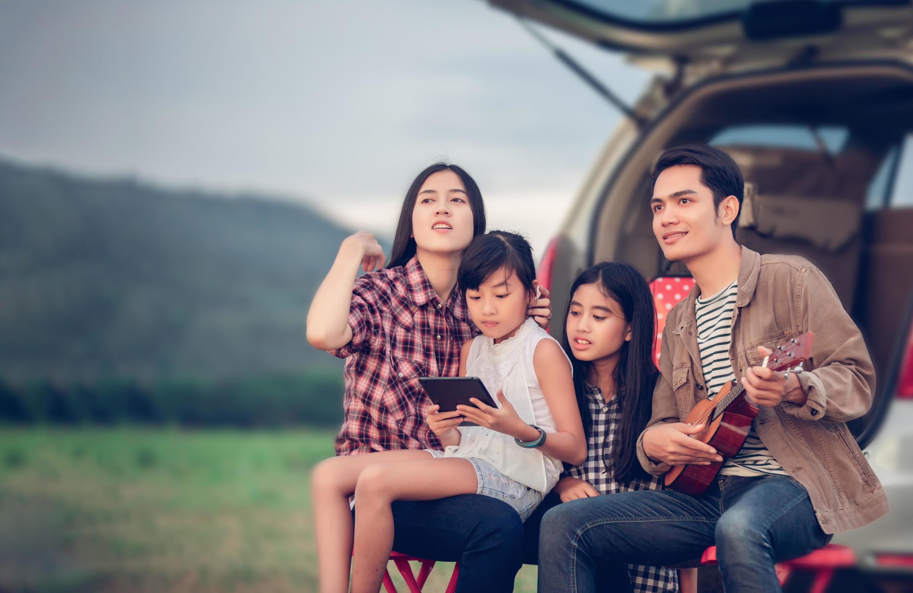 happy little girl playing ukulele with asian family sitting in the car for enjoying road trip and summer vacation Stock Free