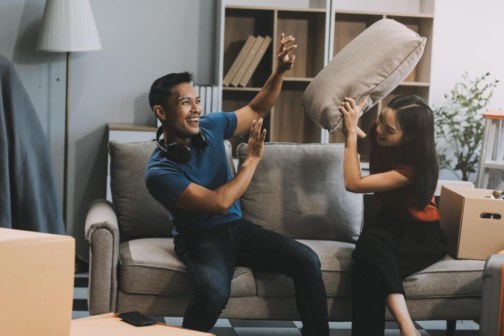 Young happy attractive Asian couple teasing each other with pillow fight on bed. White curtain background. Concept for love and happy relationship. Stock Free