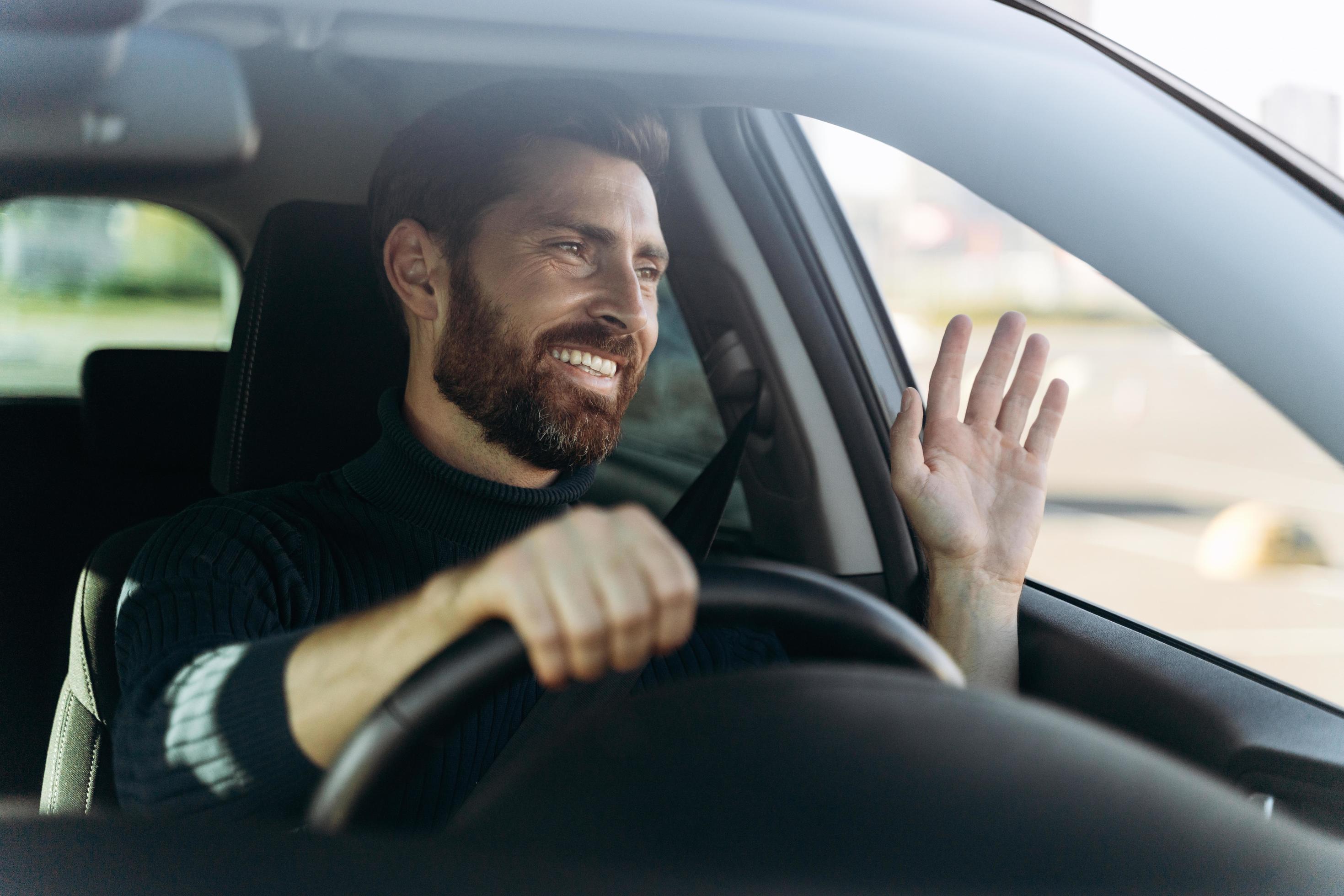 Handsome young smiling driver of car waving hand as a sign of greeting while driving the car with pleasure emotions. Transportation concept Stock Free