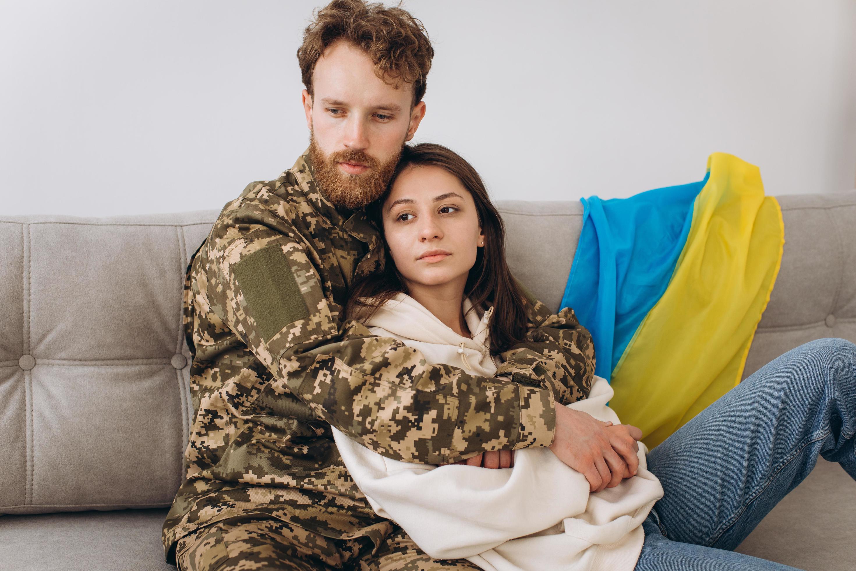 Ukrainian couple, military man in uniform with his girlfriend on the couch at home on a background of yellow and blue flag Stock Free