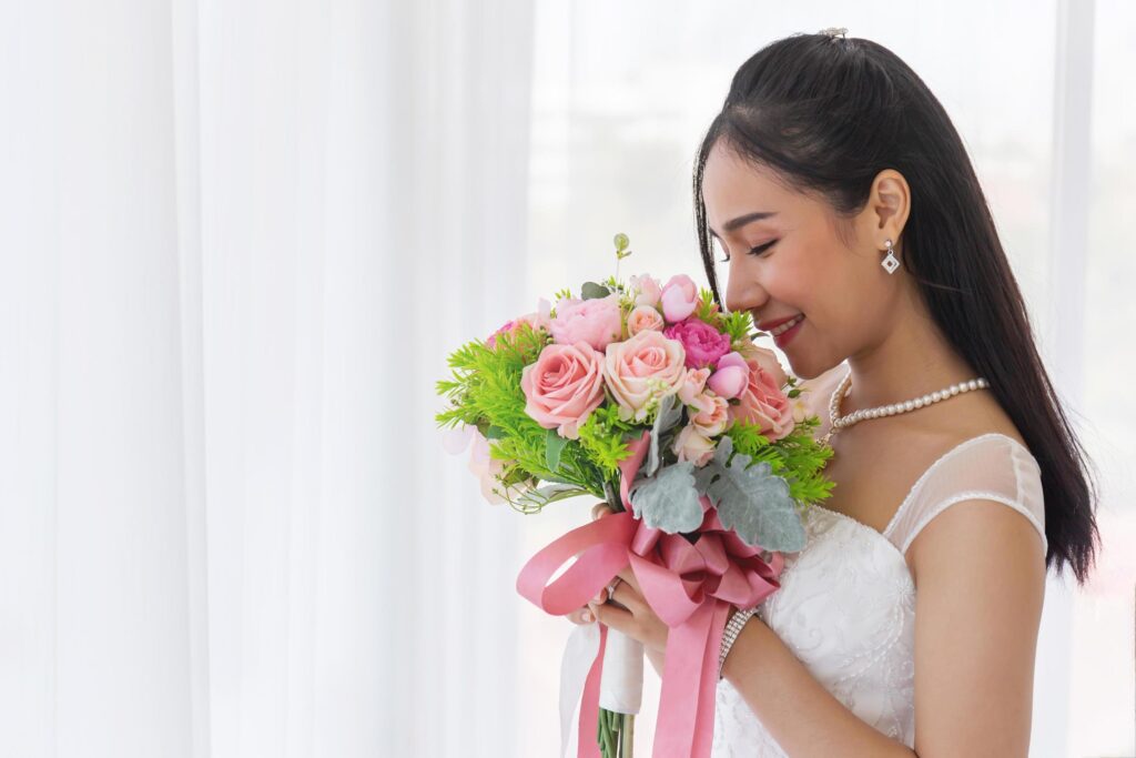 An Asian bride in a wedding dress is sitting smiling brightly on the bed in her hand holding a beautiful bouquet of flowers. Stock Free