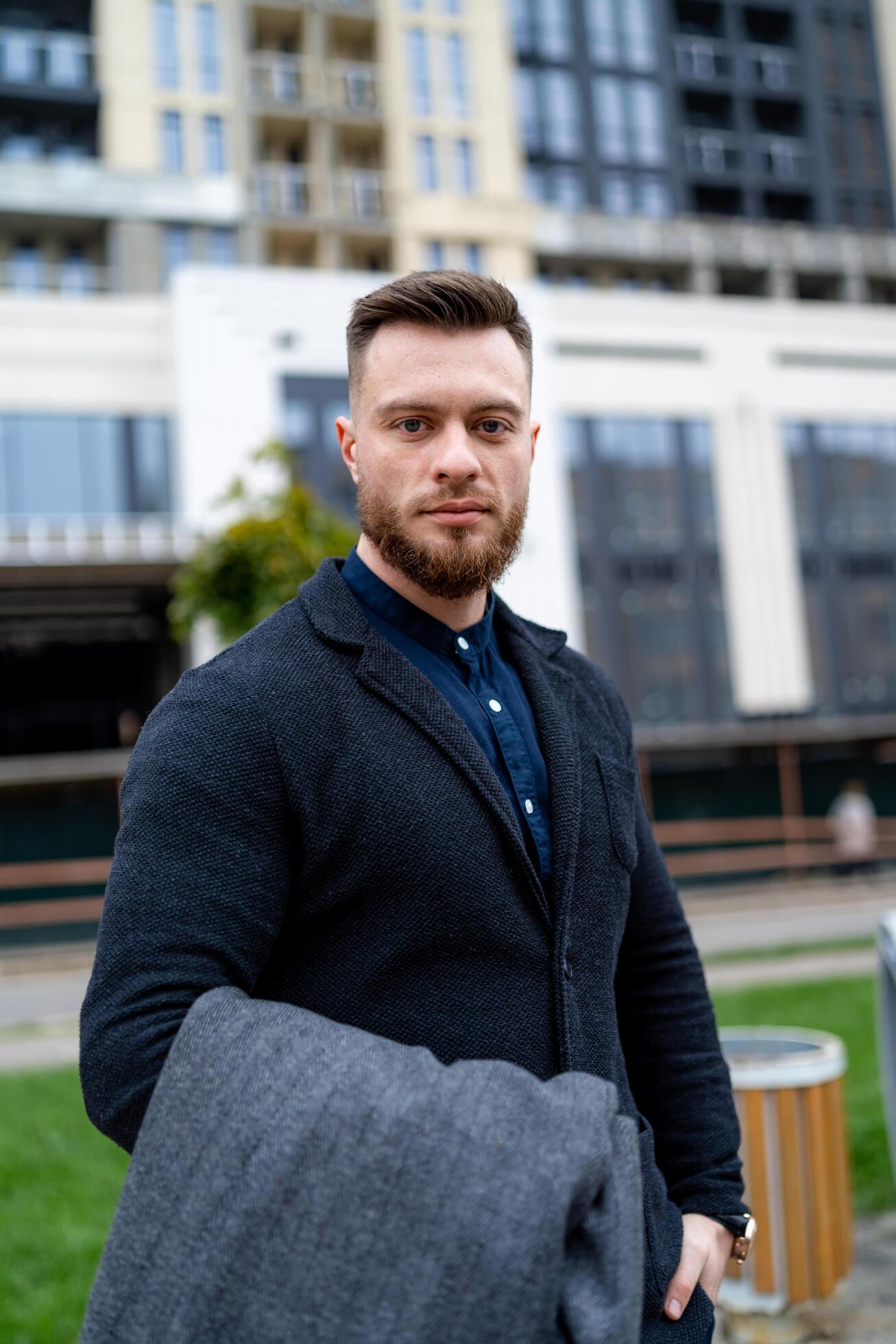 Cute man looks to the camera while wearing formal suit and holding gray coat in hands. Modern building on the background. Stock Free