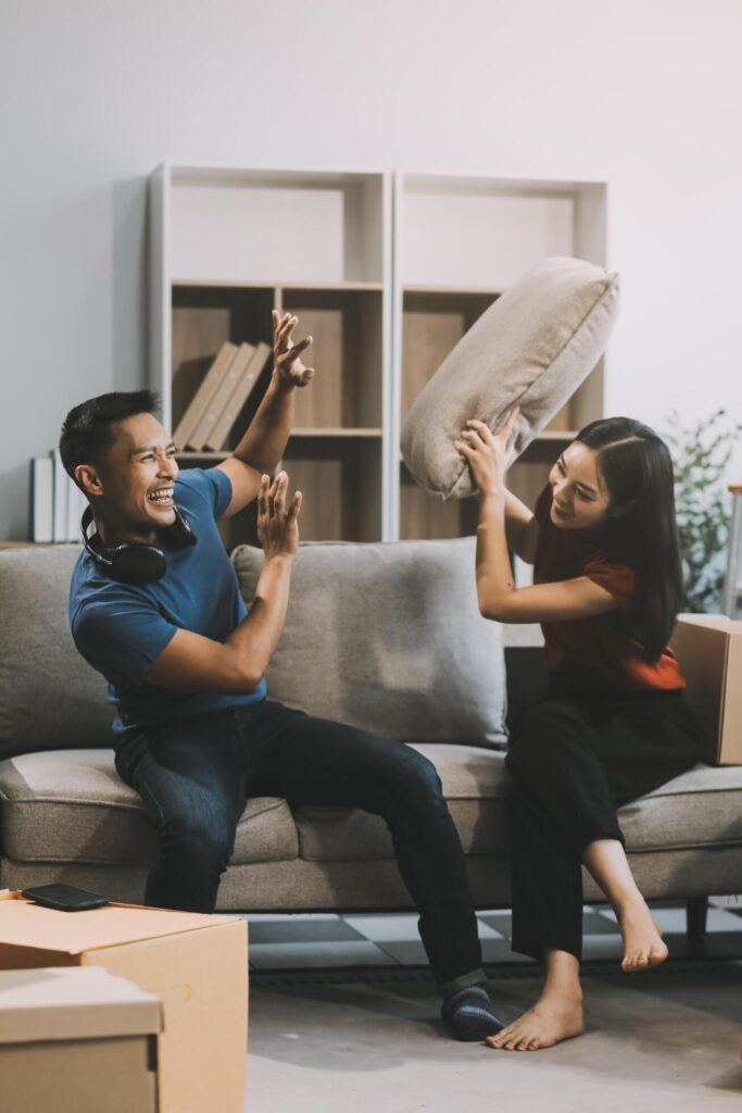 Young happy attractive Asian couple teasing each other with pillow fight on bed. White curtain background. Concept for love and happy relationship. Stock Free