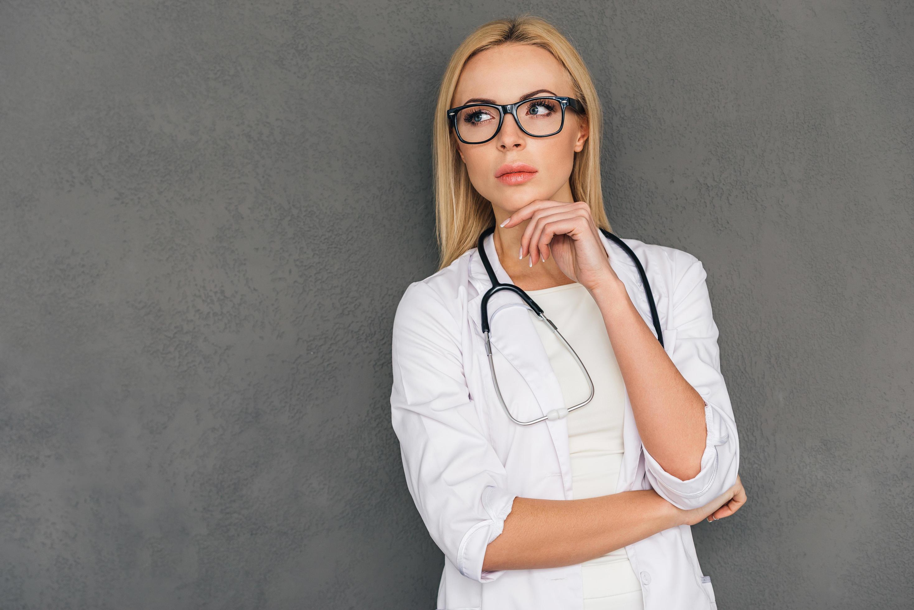 Interesting medical case. Beautiful young female doctor looking thoughtful and keeping hand on chin while standing against grey background Stock Free