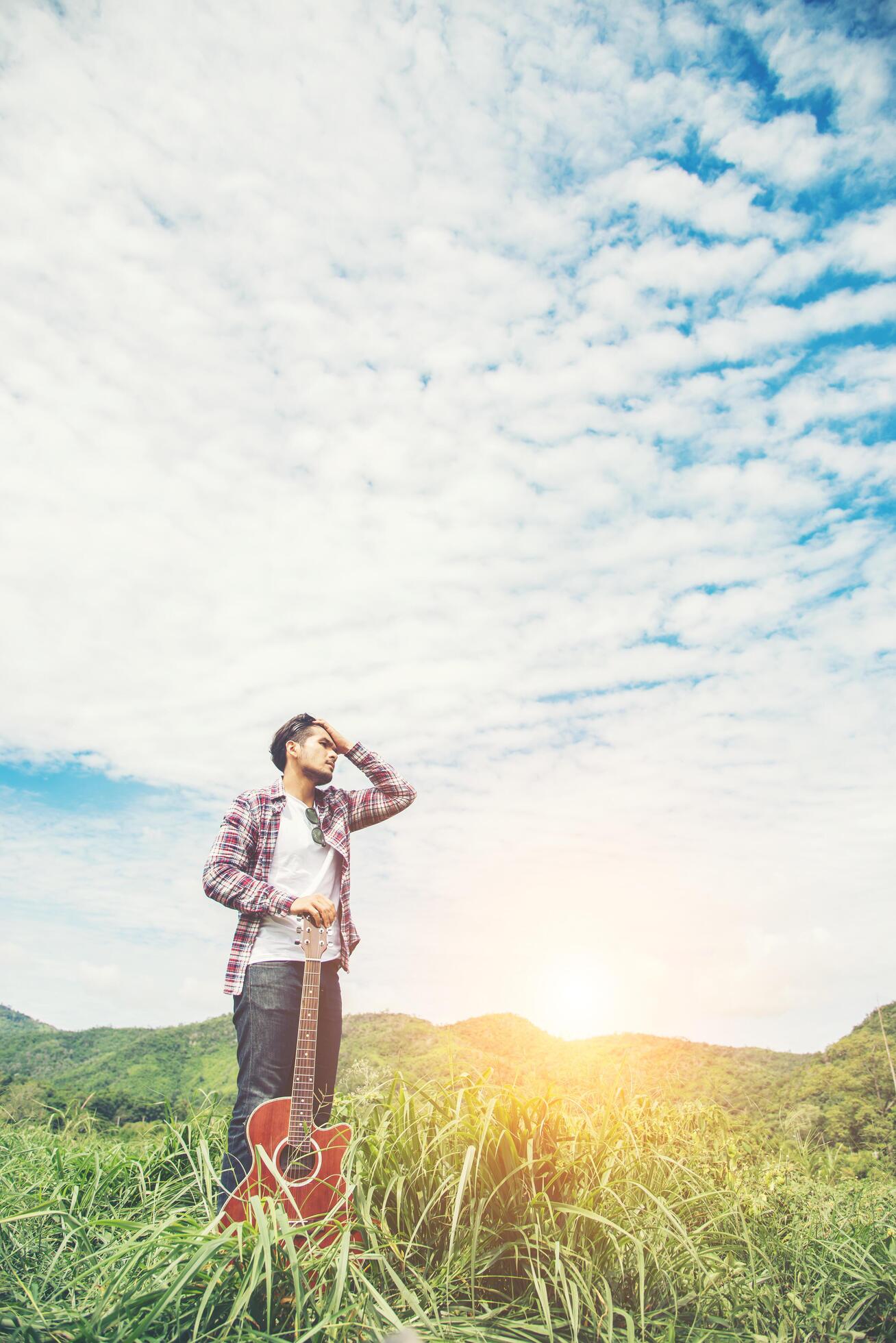 Young hipster man holding a guitar with a walking in nature, Relaxing in the field in a sunny blue sky day. Stock Free