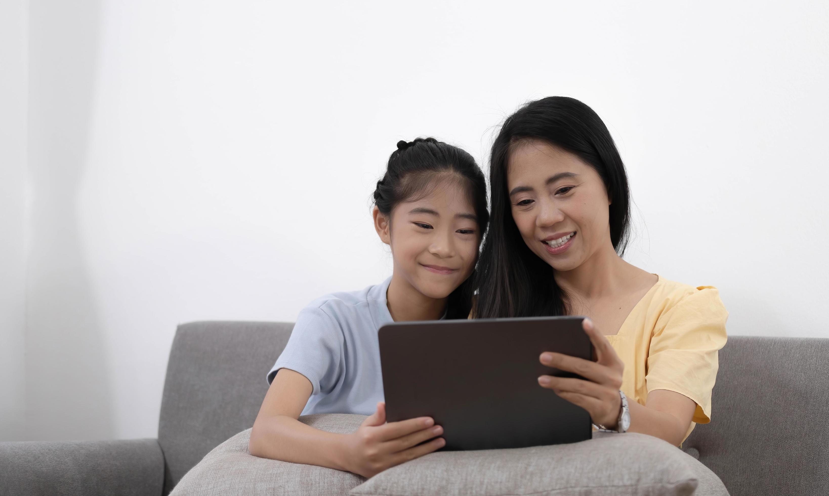 Happy young asian mom and daughter using digital tablet, watching videos or surfing internet, sitting on sofa at home, copy space. Stock Free