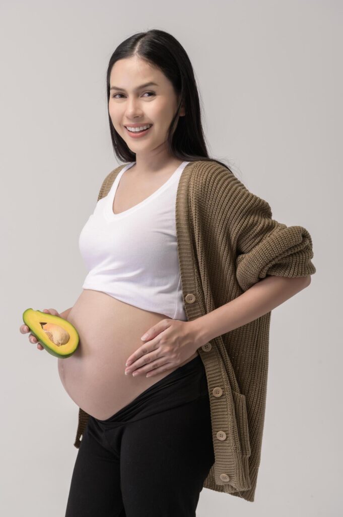 Portrait of Beautiful pregnant woman holding avocado over white background studio, health and maternity concept Stock Free