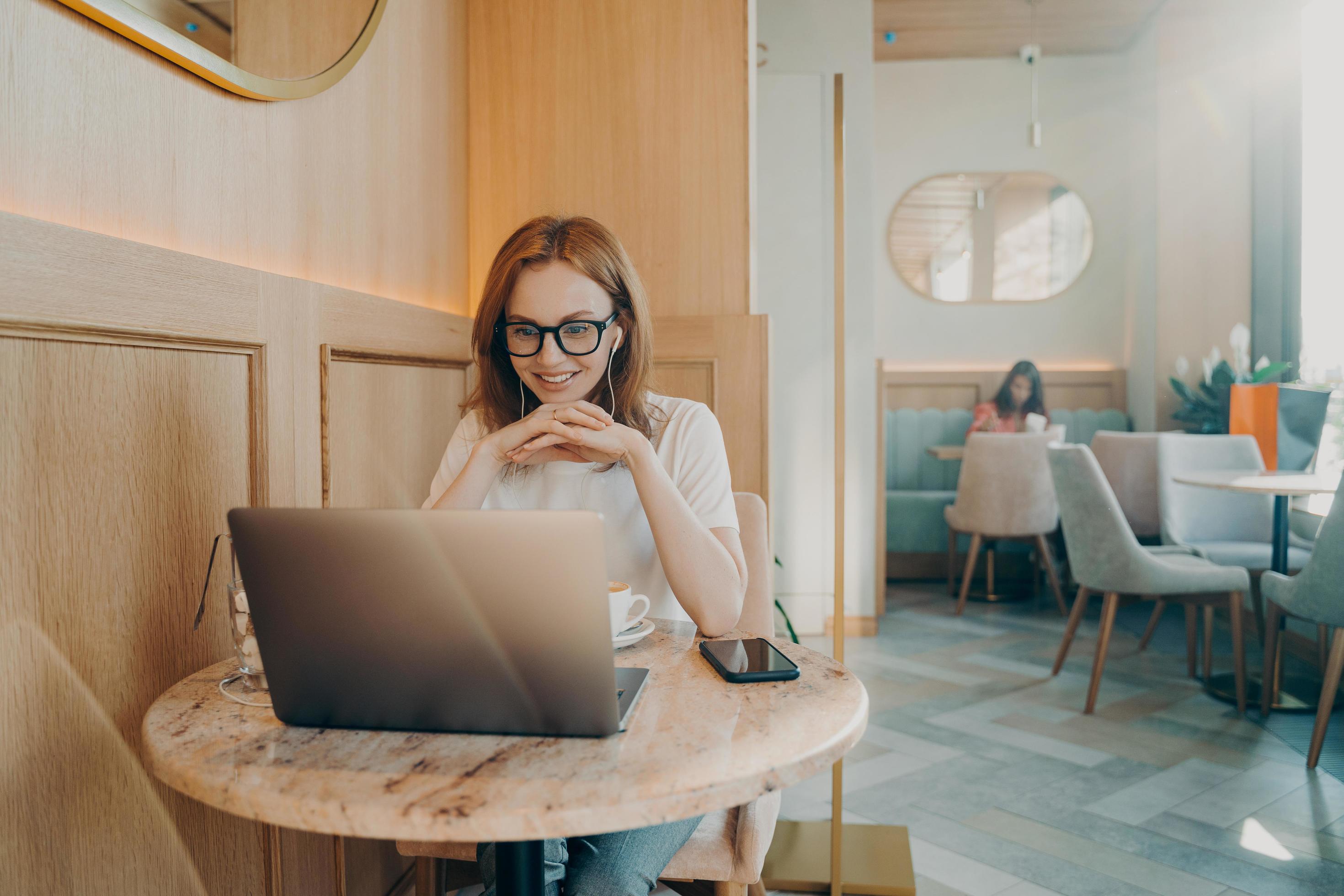 Happy redhaired woman has distance job works on laptop computer in cozy cafe Stock Free