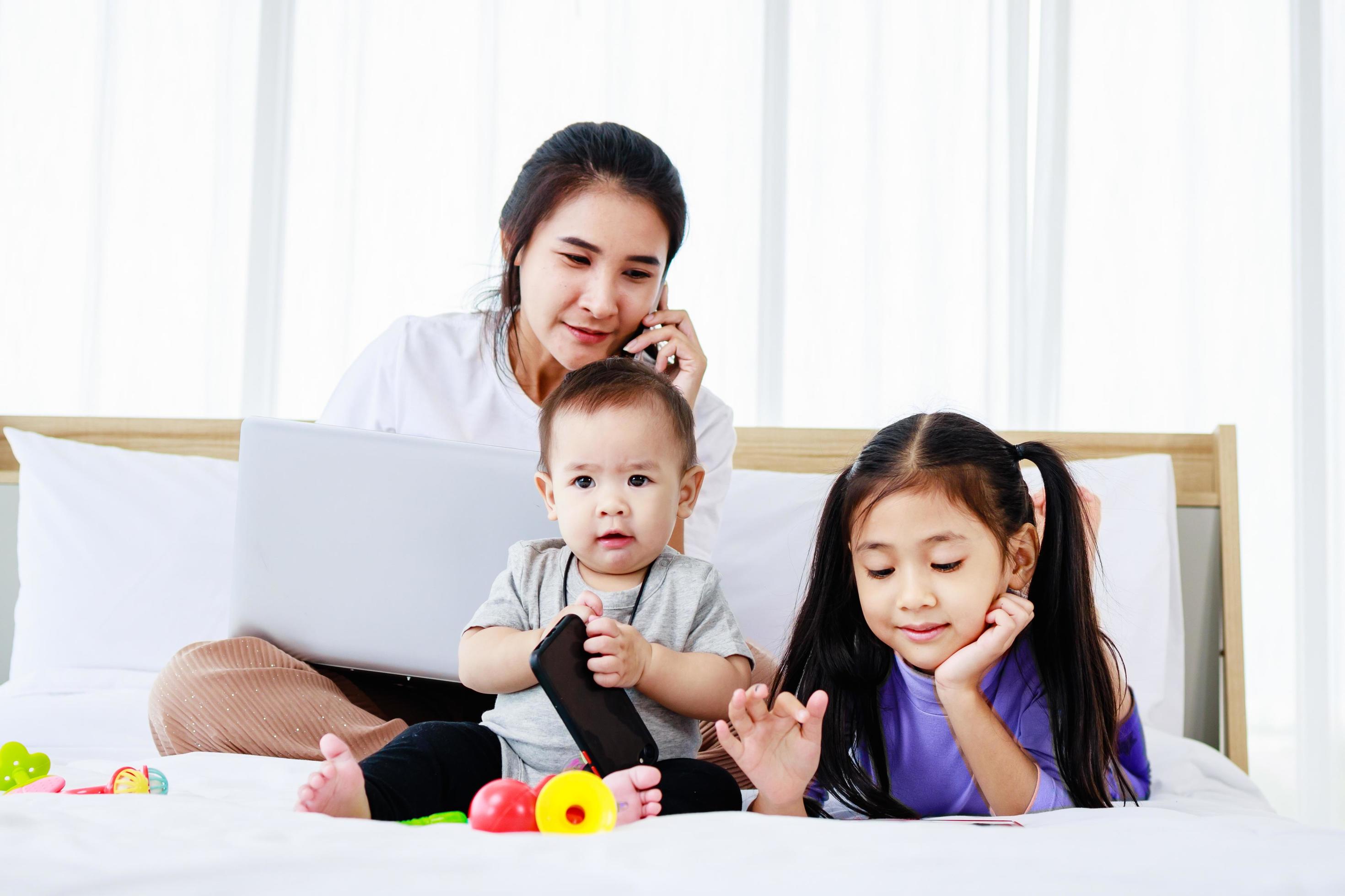 little daughter and Baby play baby toys together on bed while Busy mother working on laptop Stock Free