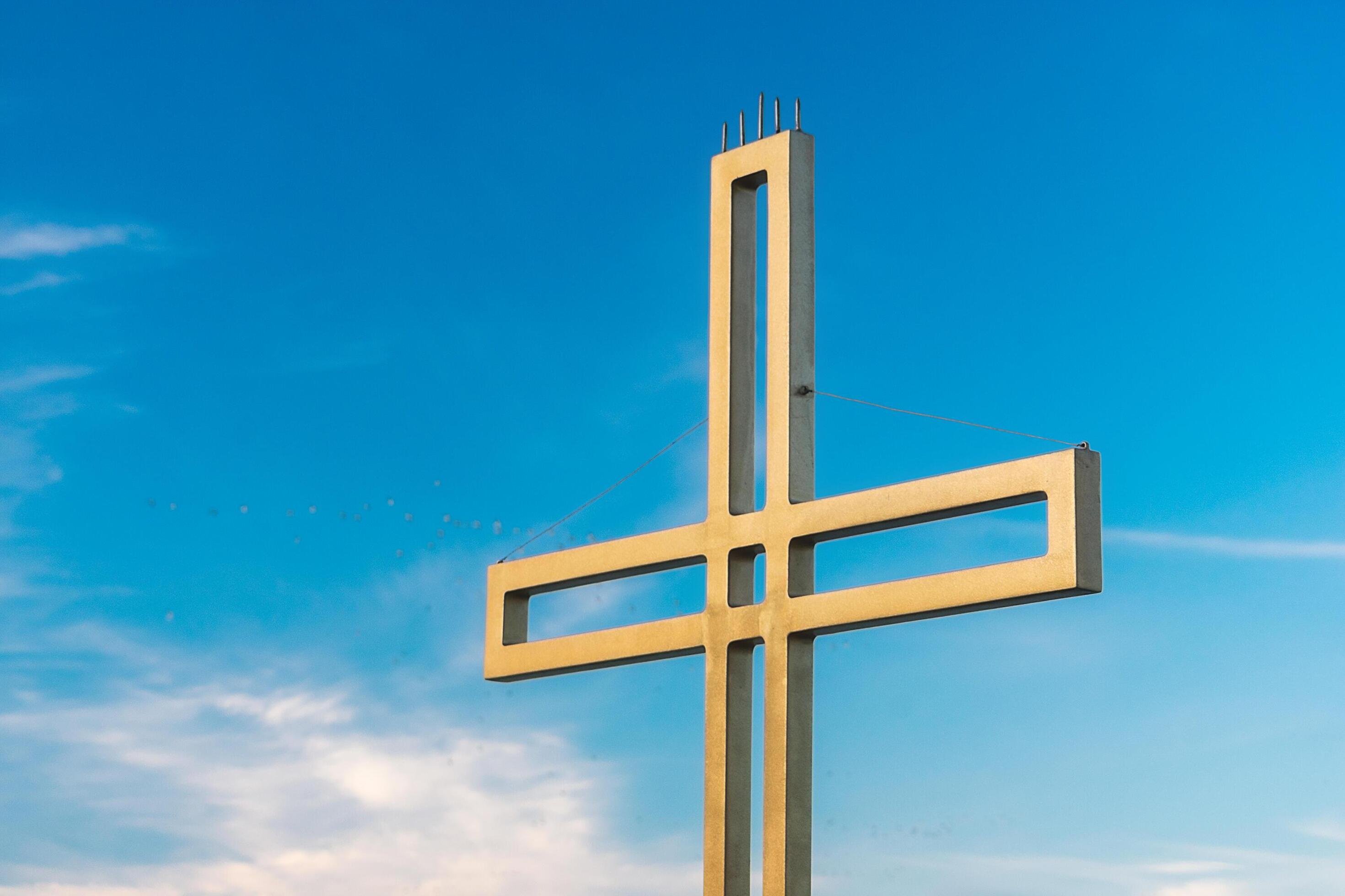 Golden cross against a background of blue sky with clouds. A minimalistic view of a gold-colored cross against the sky. Stock Free